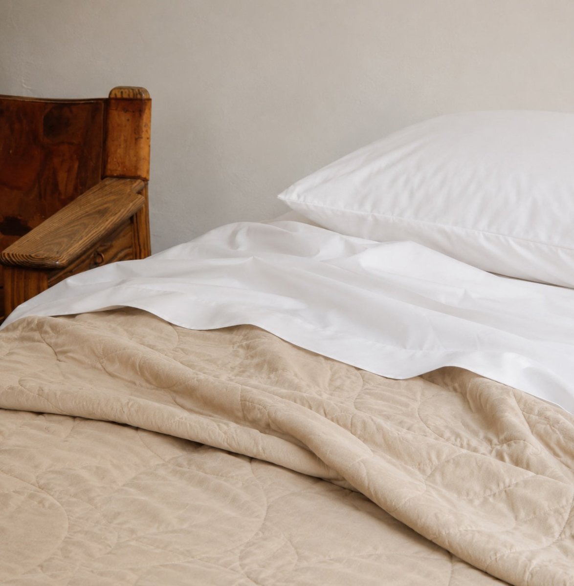 Beige quilted summer bedspread layered on white sheets with pillows on wooden bed frame